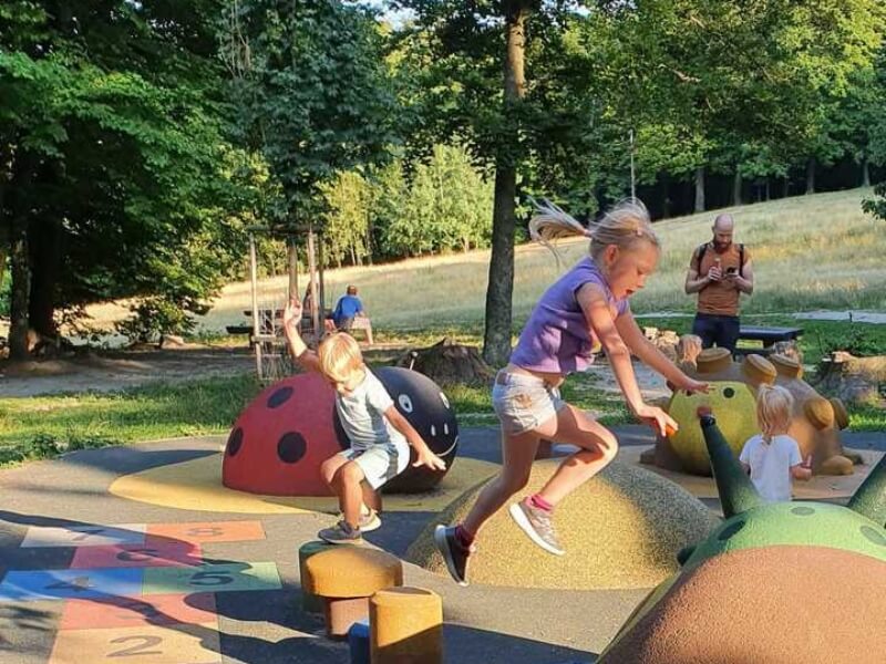 Tri deti skáču na farebnom detskom ihrisku pod korunami stromov.