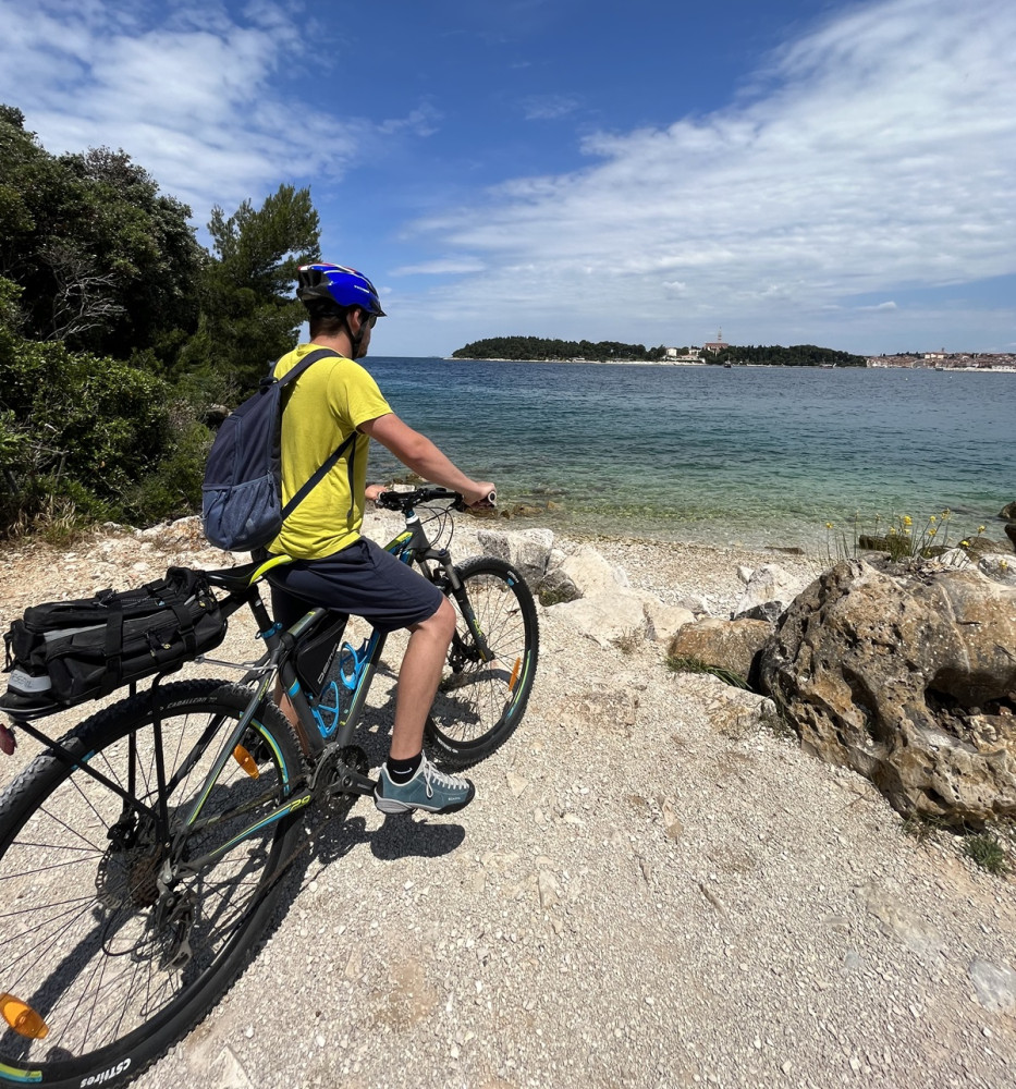 Muž na bicykli na brehu mora na štrkovej pláži s výhľadom na historické mesto.