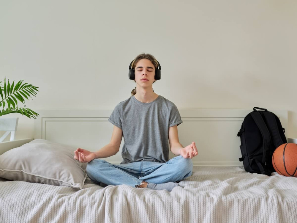 Chlapec sedí na posteli s nohami v tureckom sede a medituje.