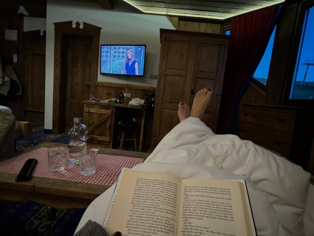 Žena s vyloženými nohami číta knihu a sleduje televízor.