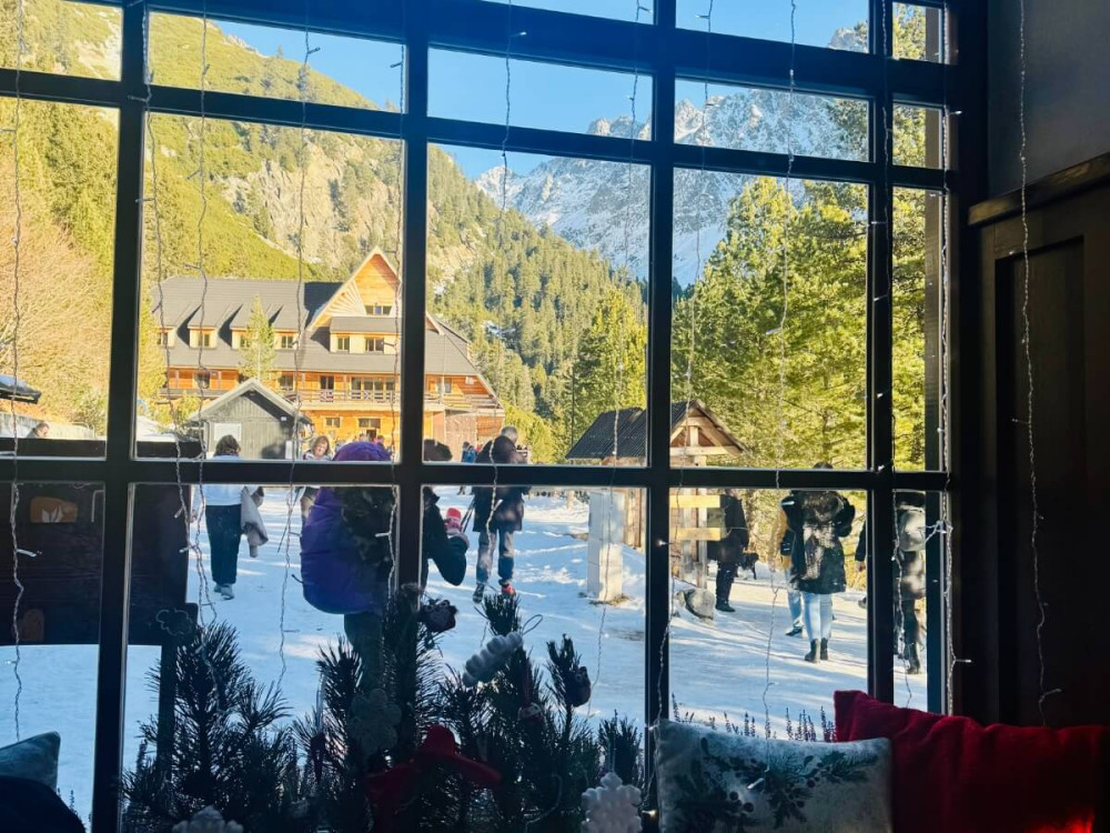 Cez okno vidieť turistov pred chatou na zasneženom chodníku.