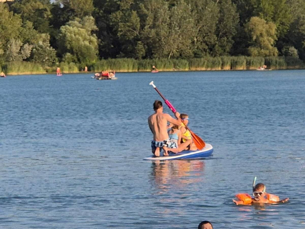 Otec so synom na paddleboarde plávajú popri chlapcovi s rukávikmi v jazere.
